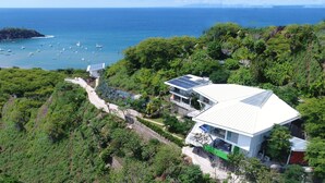 A modern house with solar panels, surrounded by lush greenery and overlooking a coastal area with boats. - The Malinche Palace (Sardinal)