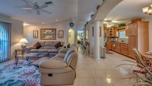 A spacious living room with a sofa set, a coffee table, and a ceiling fan. There is a kitchen area with wooden cabinets and a dining table.
