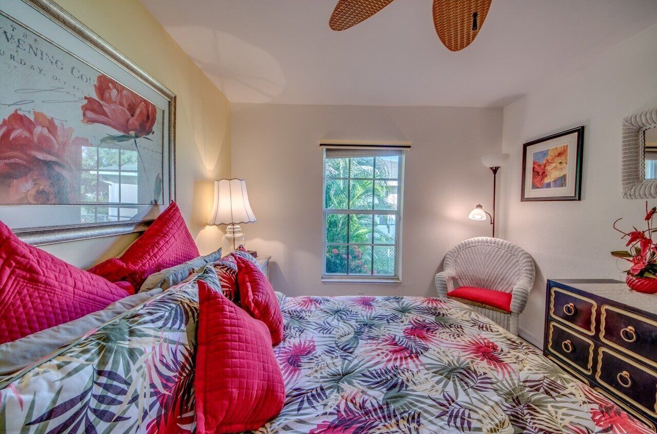 A bedroom with a floral bedspread, a large window, a wicker chair, and a dresser with drawers.