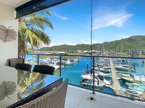 Een balkon met uitzicht op een jachthaven, palmbomen en een bergachtig landschap.