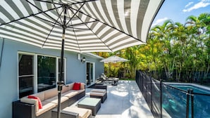 A modern outdoor patio with a pool, outdoor seating, and a large striped umbrella providing shade. - Grey Manor – Spacious 3BR/2BA Hallandale Home, Heated Pool & Smart Amenities (Hallandale Beach)