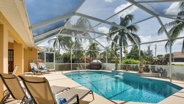 Una piscina techada al aire libre con sillas de descanso, una parrilla y palmeras al fondo.
