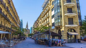 Una calle bordeada de cafés y restaurantes, con mesas al aire libre y un edificio histórico con balcones ornamentales.