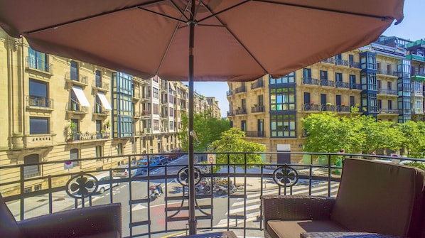 A balcony with a large umbrella, overlooking a street with parked cars and a pedestrian crossing.