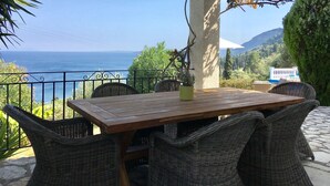 Een houten tafel met rieten stoelen, een plant in pot en uitzicht op zee en bergen.