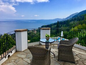 A balcony with wicker chairs, a table with a flower pot, and a view of the sea and hills. - Prestige Villas | EAST COAST | Villa with Infinity Pool & Stunning View (Corfu)