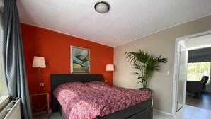 A bedroom with a bed, two bedside lamps, a painting, a potted plant, and a view to the outside. - Parkweg 21, Westkapelle (Westkapelle)