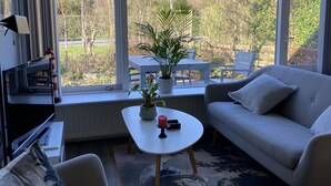 A modern living room with a grey sofa, a white coffee table, and a large window offering a view of trees and a garden. - Parkweg 21, Westkapelle (Westkapelle)