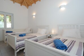 Une chambre avec trois lits simples, chacun recouvert d’une couverture à rayures et agrémenté d’une serviette bleue. On y trouve des tables de chevet blanches avec des lampes, une fenêtre avec des stores et un plafond en bois.