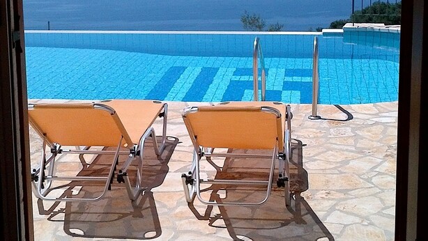 A poolside view with two sun loungers and a clear view of the sea and distant mountains. - Prestige Villas  |  NISSAKI  |  5* Stone Olive Press, Large Pool with Sea Views (Corfu)