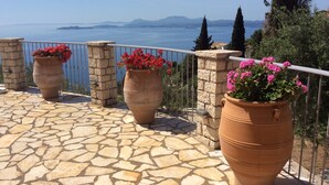A stone-tiled terrace with large terracotta pots of flowers, a stone wall, and a view of the sea and mountains. - Prestige Villas  |  NISSAKI  |  5* Stone Olive Press, Large Pool with Sea Views (Corfu)