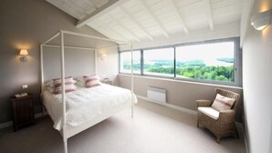 Un dormitorio con cama con dosel, vistas a zonas verdes a través de amplios ventanales, una silla de mimbre y un techo blanco.
