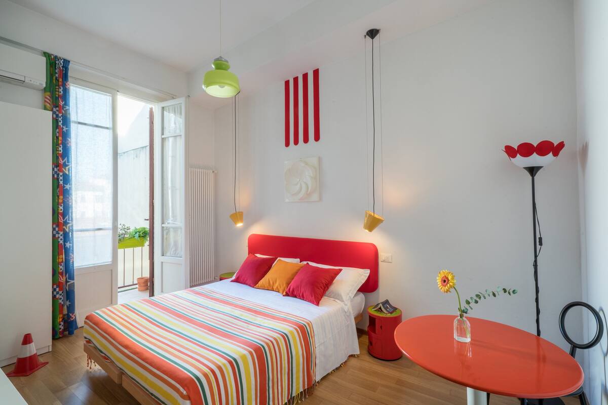 A bedroom with a bed, a red headboard, a bedside table with a vase of flowers, and a red round table.