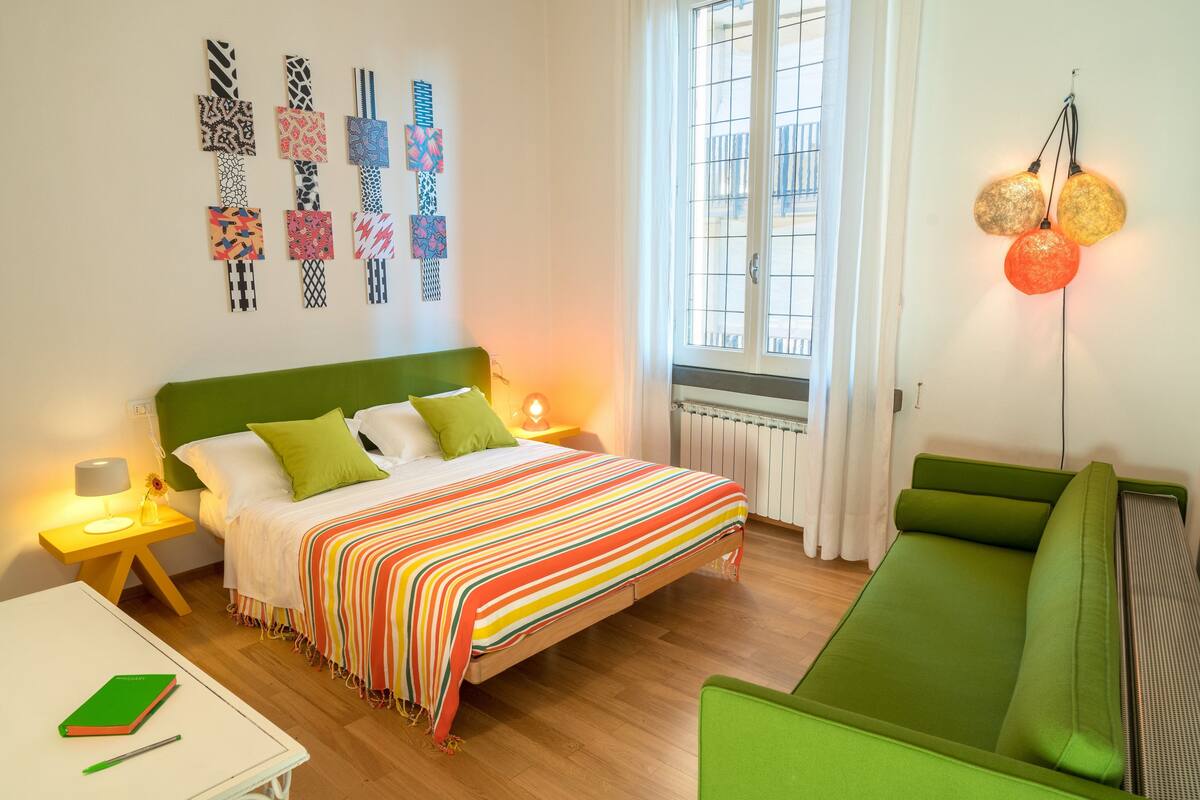 A bedroom with a bed, green sofa, bedside tables, a window, and wall art.