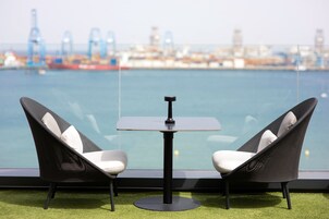 Un balcone con due sedie nere e un tavolino, affacciato su un porto con navi e gru.