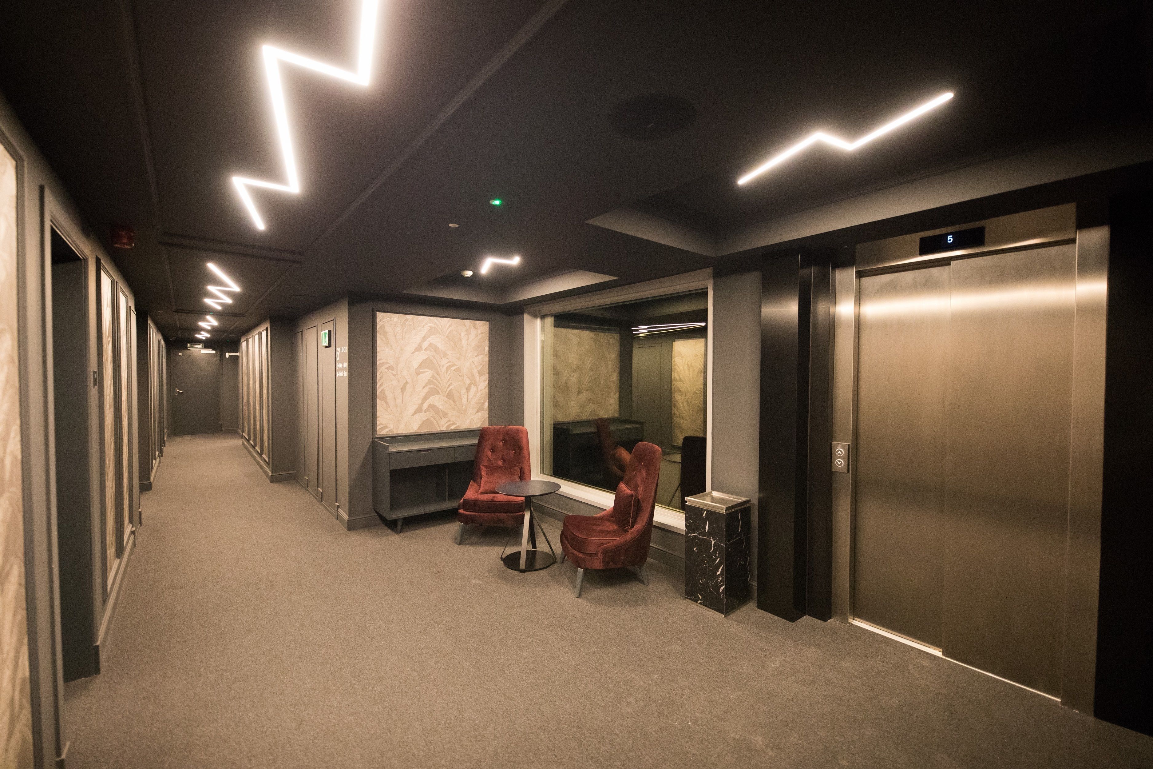 A hallway with a carpeted floor, two red chairs, a small table, and a wall with a marble-like pattern. There are two elevators, one on each side of the hallway.