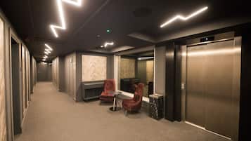A hallway with a carpeted floor, two red chairs, a small table, and a wall with a marble-like pattern. There are two elevators, one on each side of the hallway.