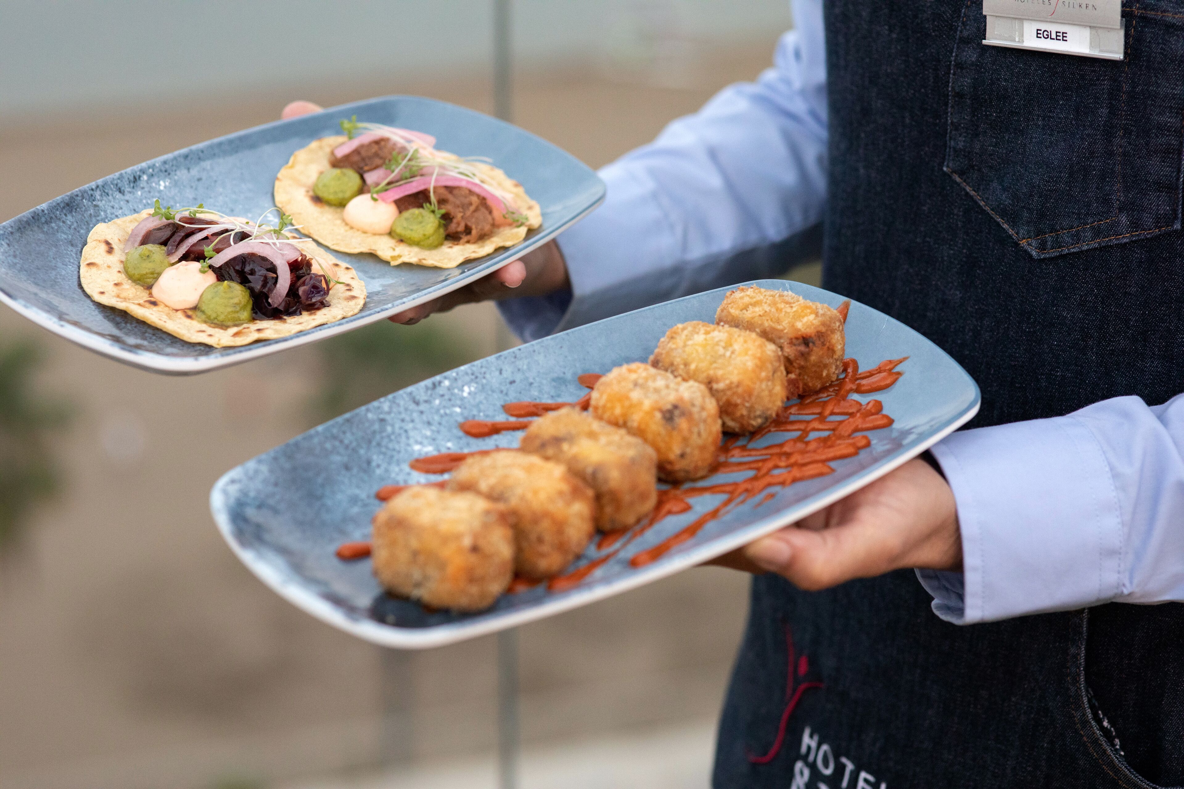 Una persona sosteniendo dos platos de comida, uno con tacos y otro con buñuelos, en un entorno al aire libre.
