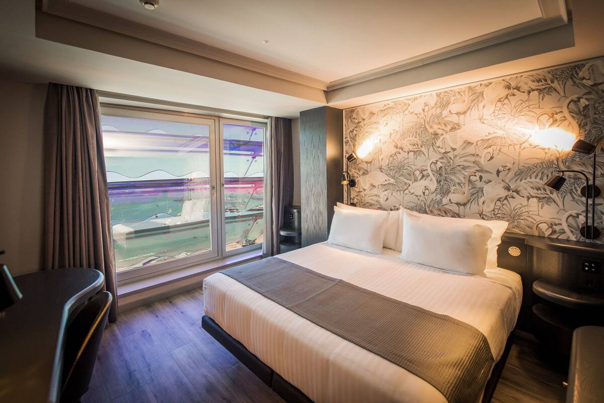 Ein Hotelzimmer mit einem großen Bett, einem Schreibtisch und Blick auf eine Stadtlandschaft durch eine Schiebetür.
