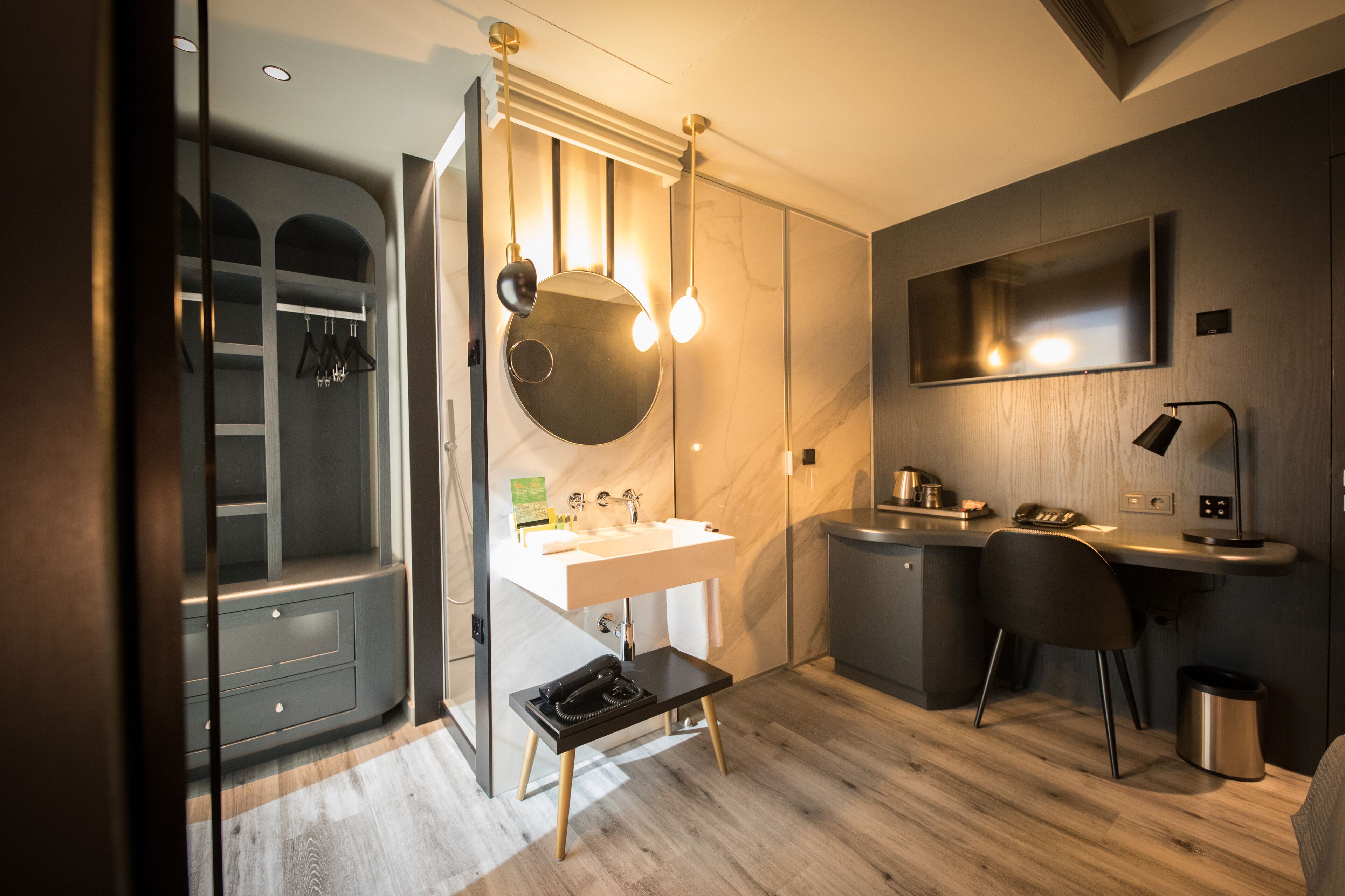 Un baño moderno con tocador, lavamanos y espejo. Hay una zona de ducha con puerta de vidrio, un perchero y un taburete pequeño.