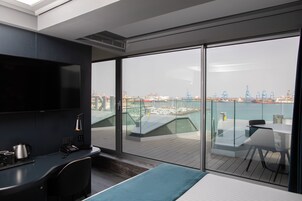 A modern hotel room with a large balcony, a desk, a bed, and a TV.