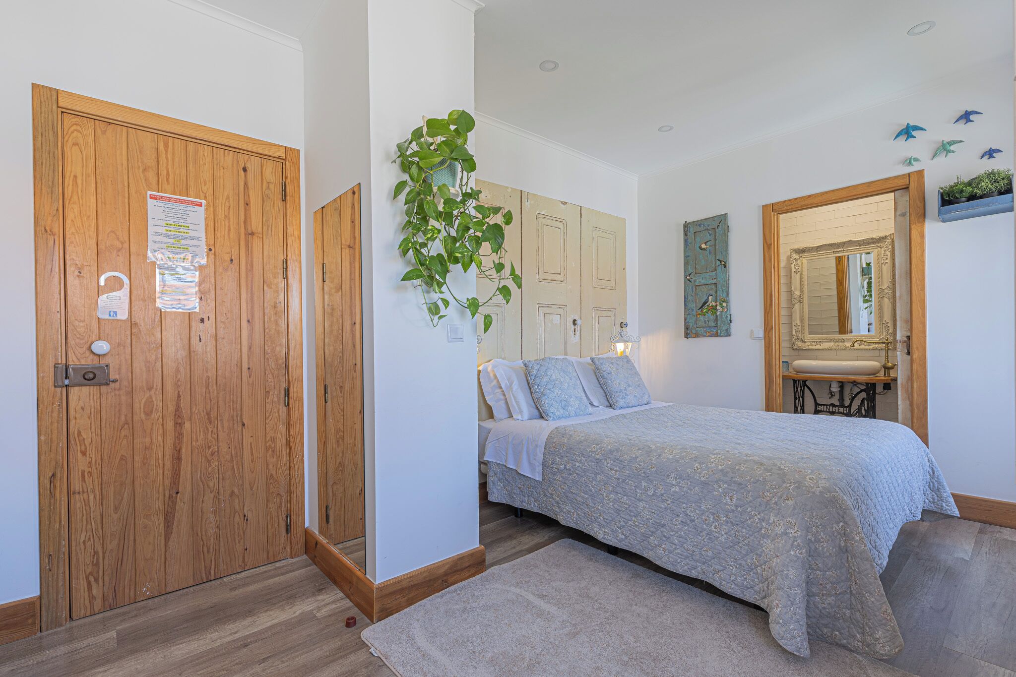 Un dormitorio con puerta de madera, una cama con colcha azul, una planta colgante y un espejo con marco decorativo.