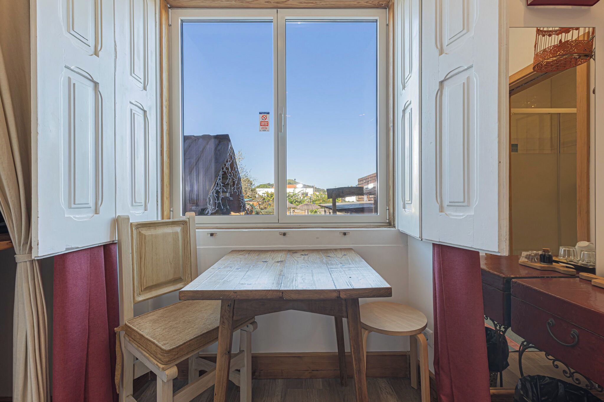 A room with a wooden table and two chairs, a window with a view of buildings, and a red curtain.