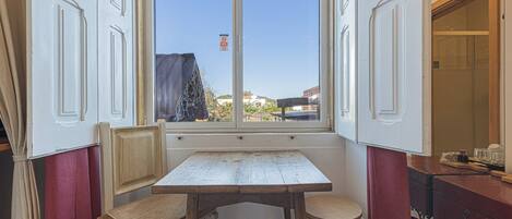 A room with a wooden table and two chairs, a window with a view of buildings, and a red curtain.