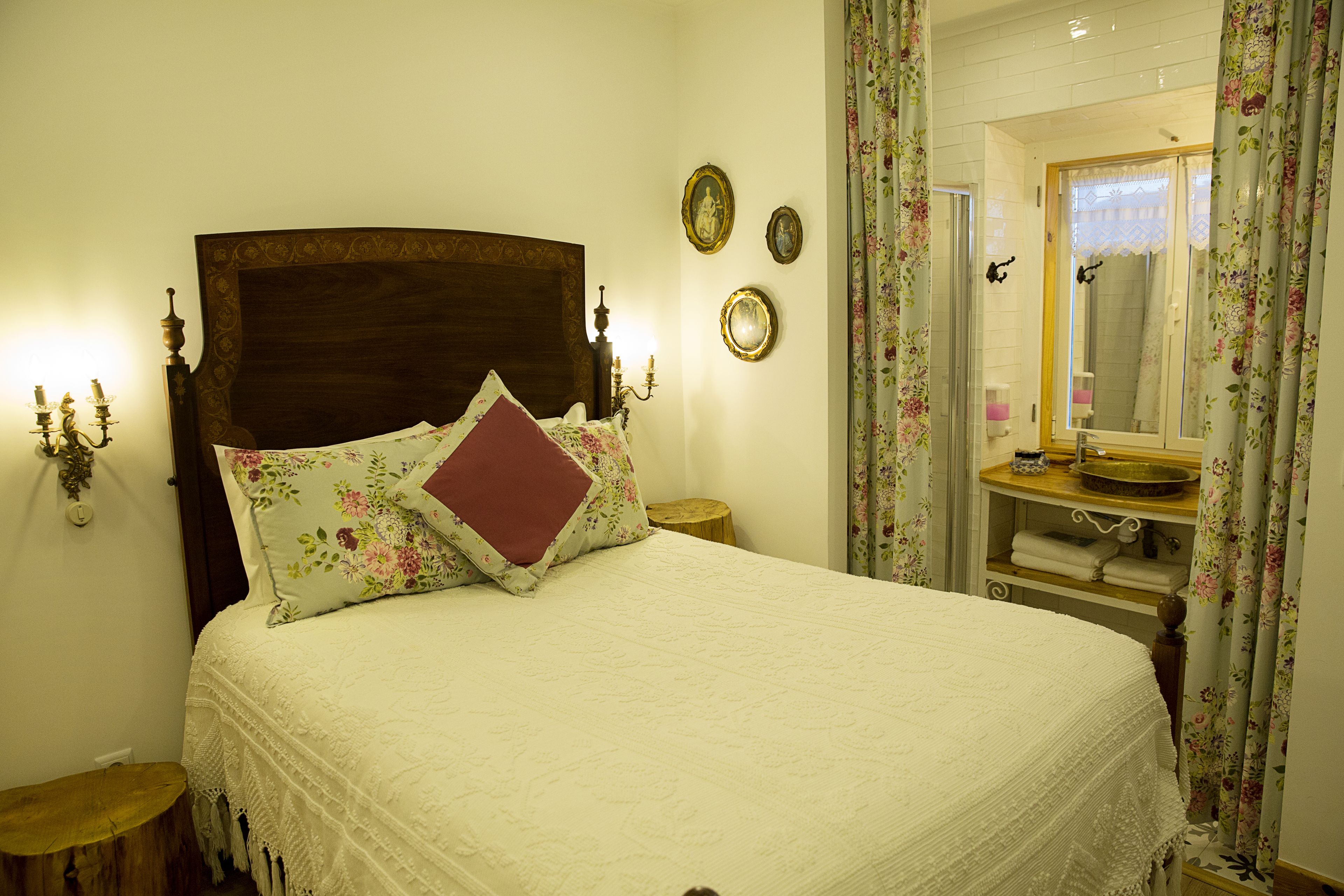Una cama bien tendida con cabecera de madera, una mesita de noche con lámpara, y un baño con espejo y lavabo.