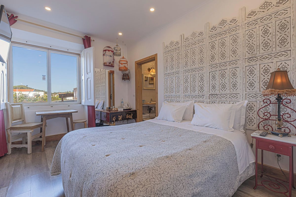 Ein Schlafzimmer mit einem großen Bett, einem Schreibtisch und einem Stuhl. Ein Fenster mit Vorhängen, eine Wand mit einem dekorativen Muster und eine Lampe auf einem Nachttisch.