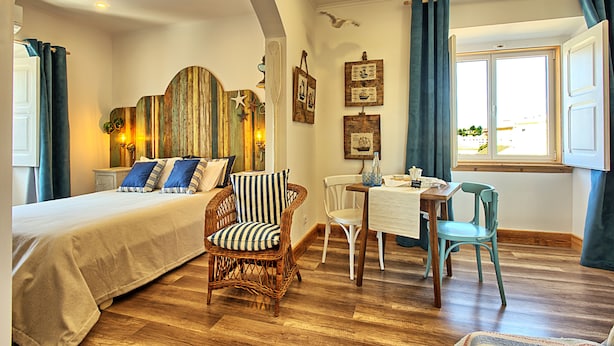 Une chambre à coucher avec un lit, une chaise en osier, une table à manger avec des chaises et une fenêtre avec des rideaux.