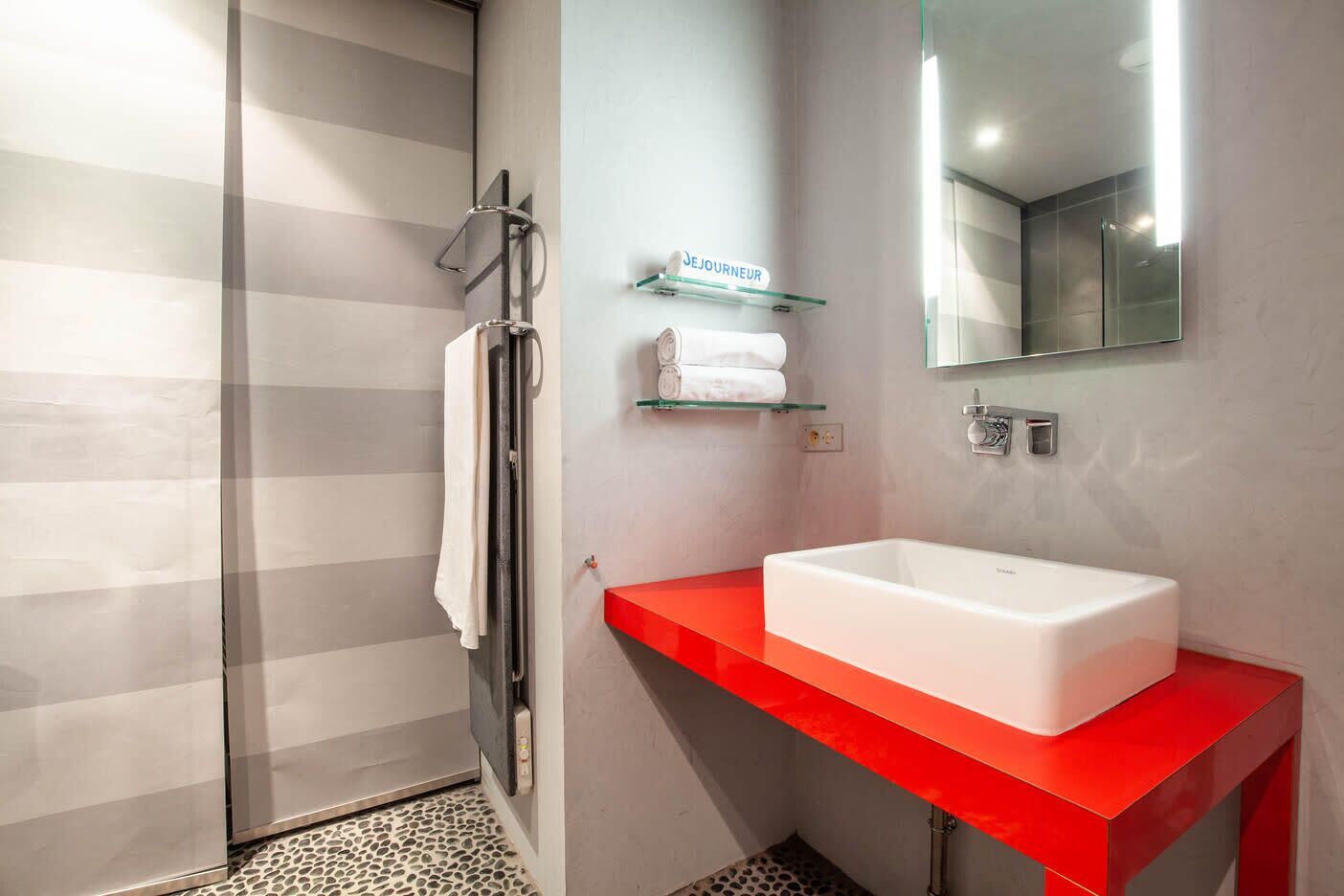 Un baño moderno con cabina de ducha de vidrio, un lavamanos blanco sobre una encimera roja y un espejo por encima.