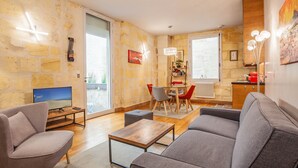 A modern living room with a sofa, armchair, coffee table, and a TV. There's a dining area with a table and chairs, and a kitchen area with a radiator and a window. - Beautiful stone flat for 4 in the Rue des Antiquaires (Bordeaux)