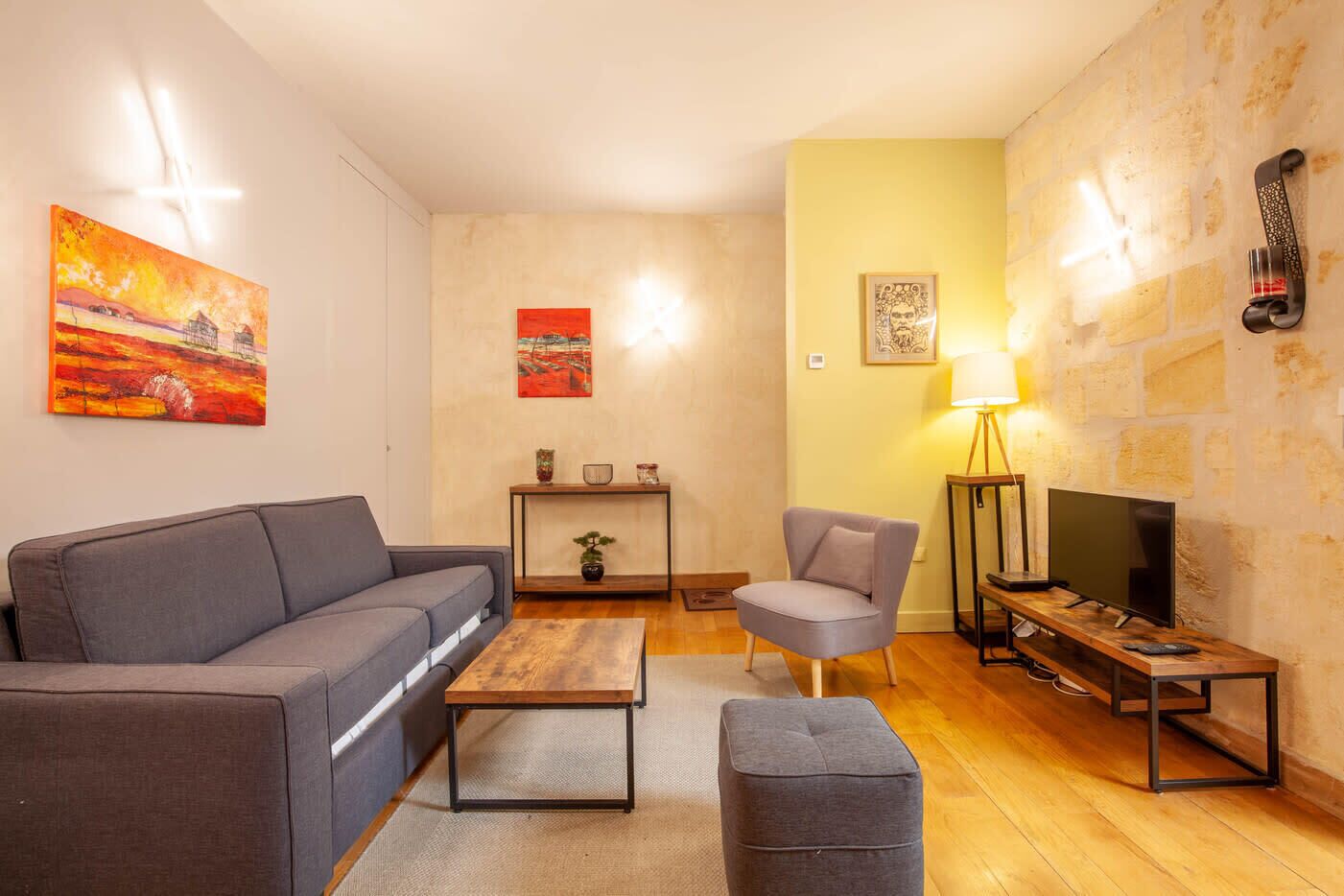 Ein Wohnzimmer mit Sofa, Sessel, Couchtisch und Hocker. Es gibt einen Fernseher, eine Lampe und Kunstwerke an den Wänden.