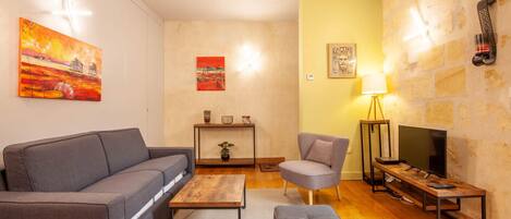 Ein Wohnzimmer mit Sofa, Sessel, Couchtisch und Hocker. Es gibt einen Fernseher, eine Lampe und Kunstwerke an den Wänden.