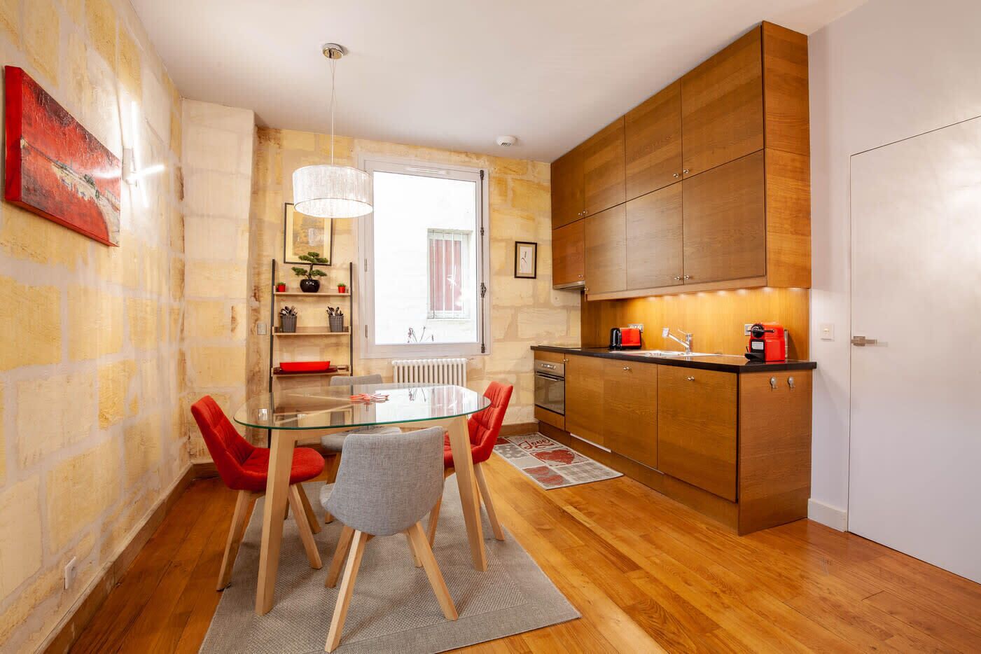 A compact kitchen with wooden cabinets, a glass dining table with red chairs, and a window providing natural light.