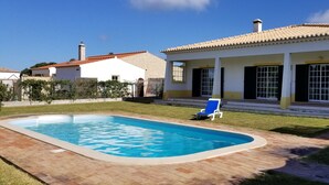 Una piscina con un borde de baldosas, una silla de descanso azul y una casa con techo de tejas y detalles en amarillo.
