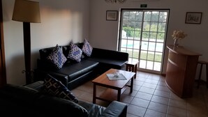 Un salón con un sofá negro, cojines estampados, una mesa de centro de madera, una lámpara y vista a un jardín por una puerta de cristal.