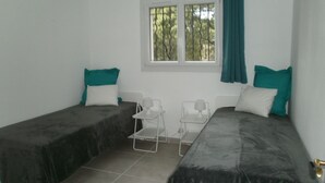 A room with two beds, each with a dark cover and teal pillows, a small table with a lamp, and a window with a green curtain. - Air cond. panoramic view, 12x6 swimming pool, diving board, on 2 hectares, quiet (Lorgues)