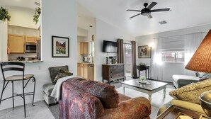 A modern living room with a sofa, coffee table, and a television. There is a kitchen area with cabinets and a microwave.