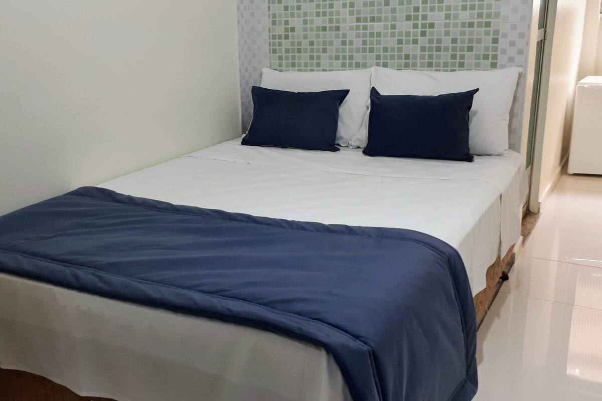 A single bed with a white sheet and a blue blanket, two dark pillows, and a white headboard with a green mosaic pattern.