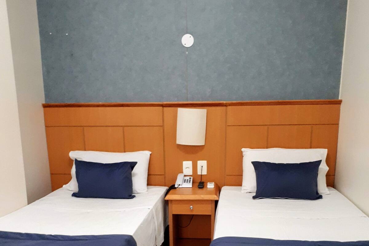 A hotel room with two beds, each with a blue bedspread and white pillows, a wooden headboard, a nightstand with a phone and a lamp, and a wall-mounted light fixture.