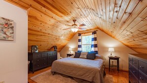 A bedroom with a sloped ceiling, a bed with a beige comforter, a wooden dresser, a nightstand with a lamp, and a ceiling fan. - Woodsy Retreat w/ Fireplace, Hot Tub & Fire Pit (McHenry)