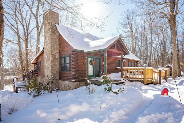 A log cabin with a stone chimney, wooden deck, and a red mailbox.