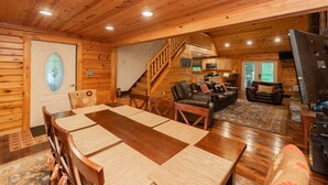 A wooden cabin interior with a dining area, living room, kitchen, and a staircase. - Woodsy Retreat w/ Fireplace, Hot Tub & Fire Pit (McHenry)