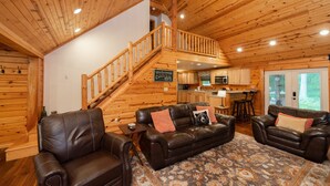 A cabin interior with a wooden staircase, leather sofas, a kitchen area, and a view of the outdoors. - Woodsy Retreat w/ Fireplace, Hot Tub & Fire Pit (McHenry)