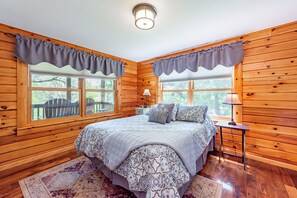 Un dormitorio de cabaña de madera acogedor, con una cama grande, dos lámparas de noche y vistas al exterior a través de dos ventanas.