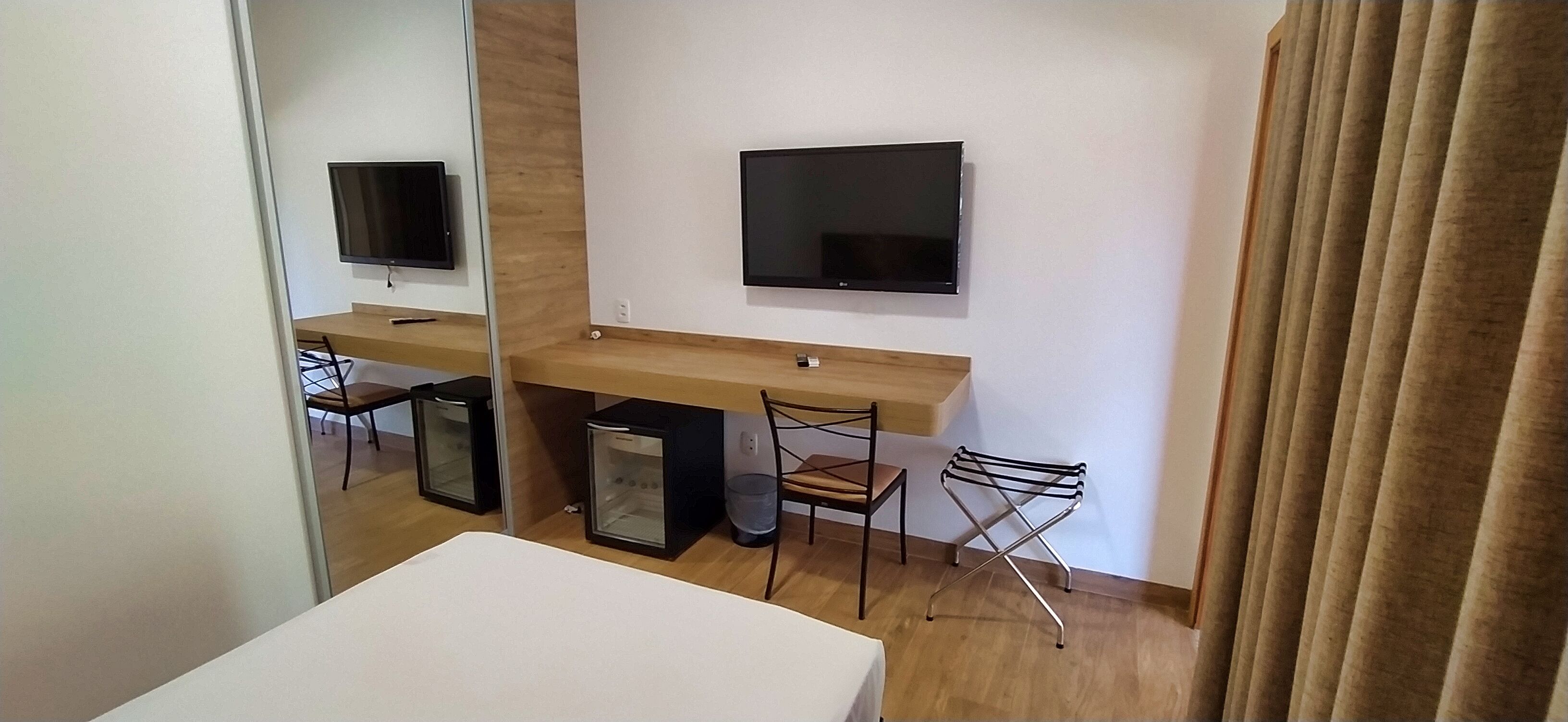 A hotel room with a wooden desk, a flat-screen TV, a chair, a small refrigerator, and a mirror.