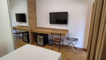 A hotel room with a wooden desk, a flat-screen TV, a chair, a small refrigerator, and a mirror.