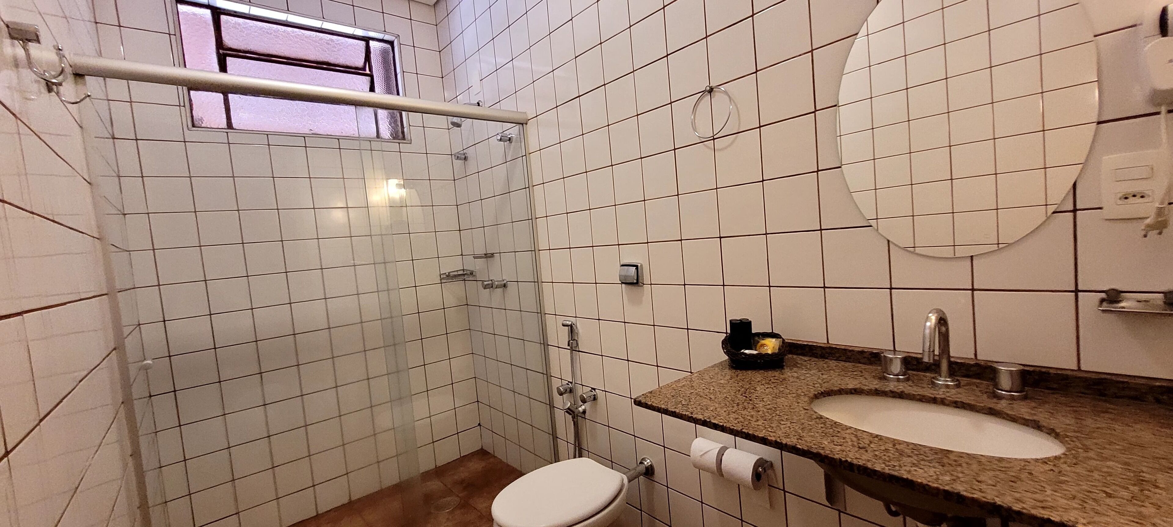 A small bathroom with white tiled walls, a shower, a toilet, and a sink with a granite countertop.